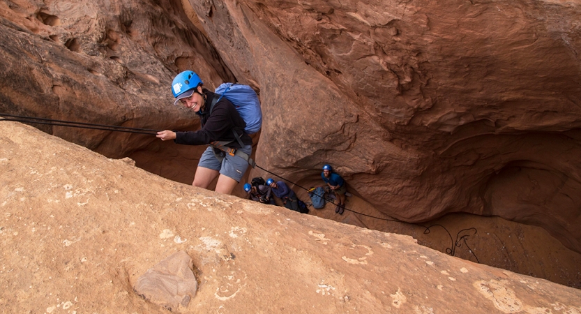 A person wearing safety gear is secured by ropes as they descend into a canyon. They are looking up at the camera and smiling.